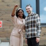 Young couple celebrates buying their first house with keys in hand and a sold sign.
